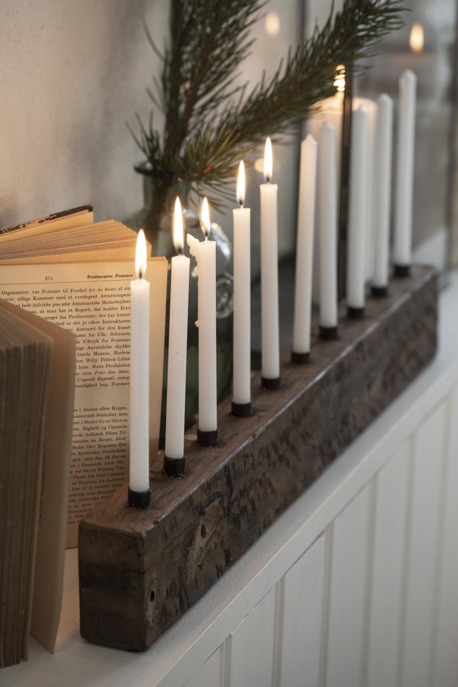 Mehrere weiße dünne Kerzen von Ib Laursen in Holzleiste als Adventsdekoration mit Buch und Tannenzweig