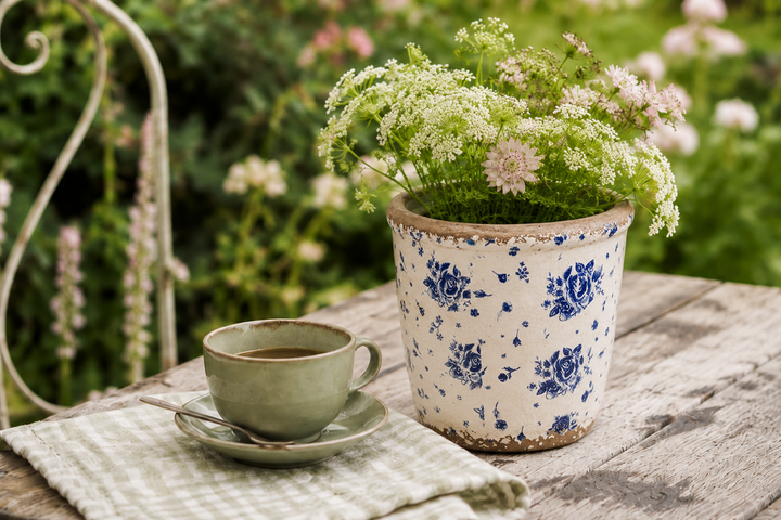Clayre & Eef Übertopf mit blauem Rosendekor und Wildblumen neben Kaffeetasse auf rustikalem Gartentisch im Shabby Chic Stil